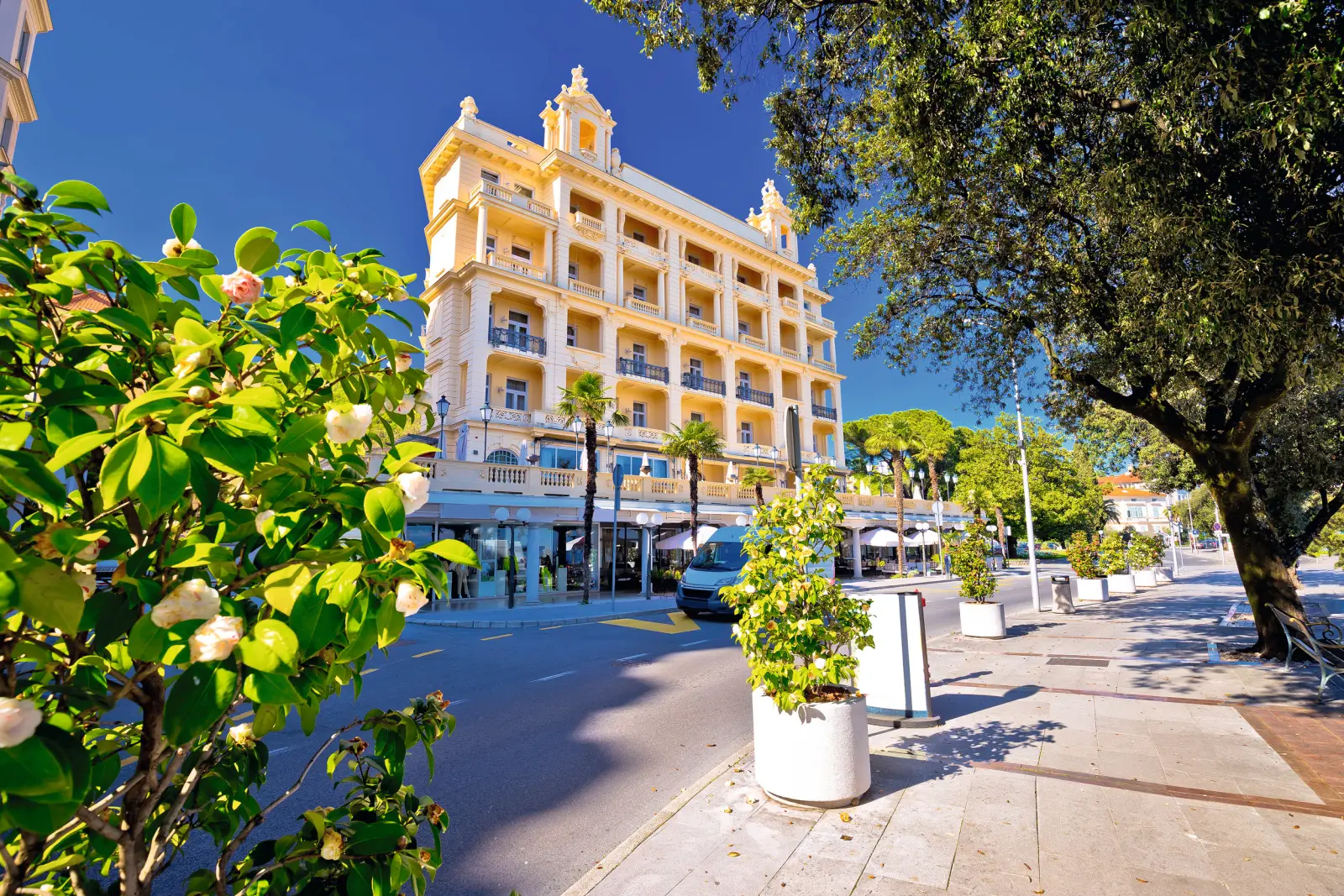Hotel giallo con balconi lungo una strada alberata sotto un cielo azzurro.