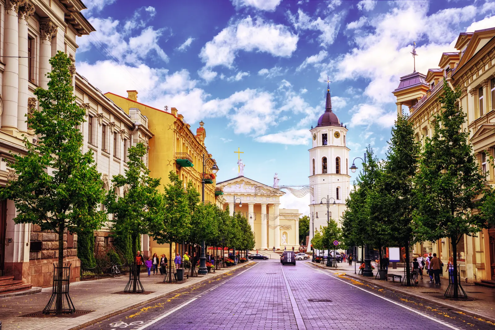 Street with trees, buildings and a church tower under a blue sky.