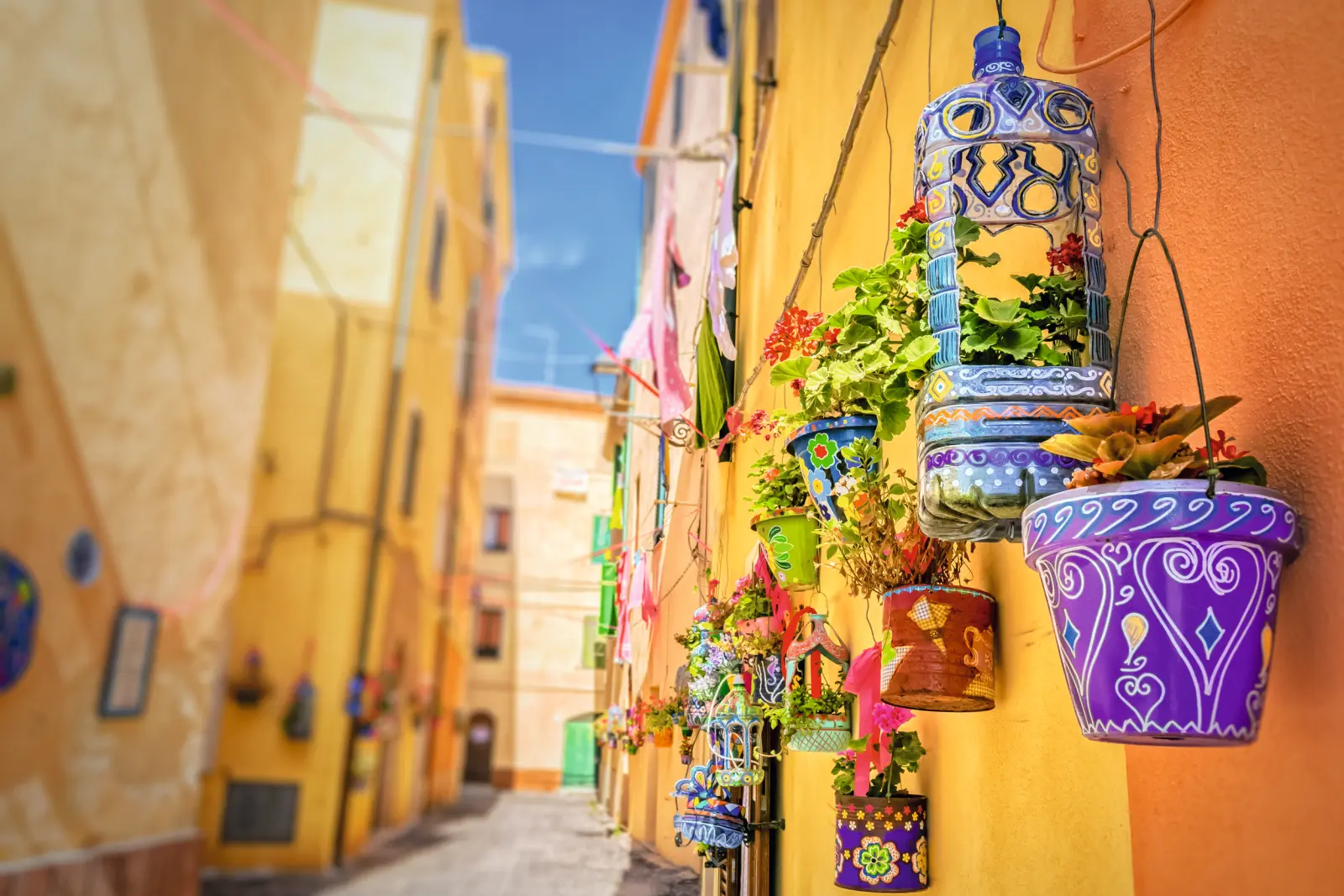 Pots de fleurs colorés contre un mur jaune dans une ruelle étroite et ensoleillée.