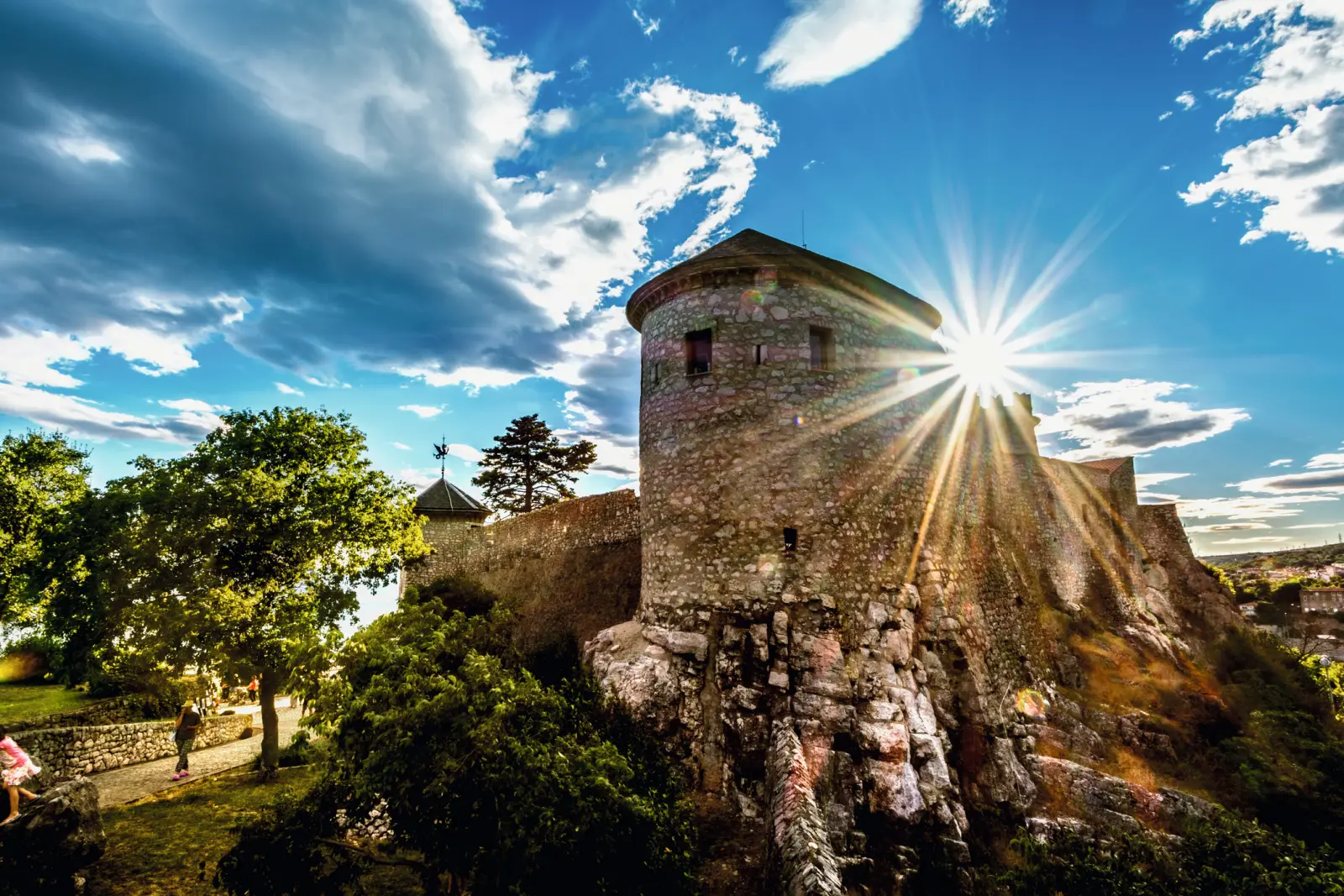 Antica fortezza in pietra con raggi di sole, circondata da alberi e cielo azzurro.