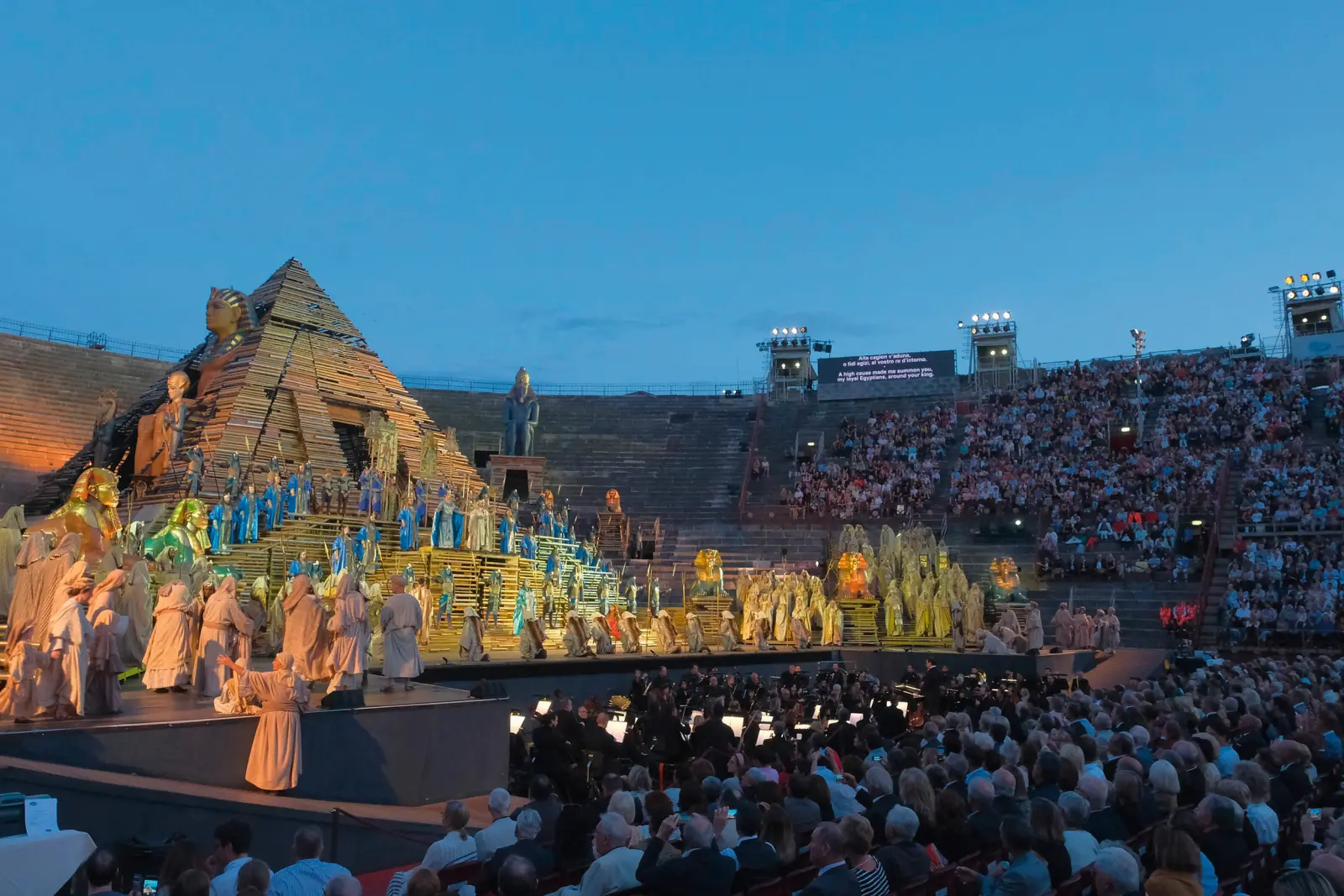 Stage performance of Verdi's Aida in an ancient amphitheatre with a pyramid and audience.