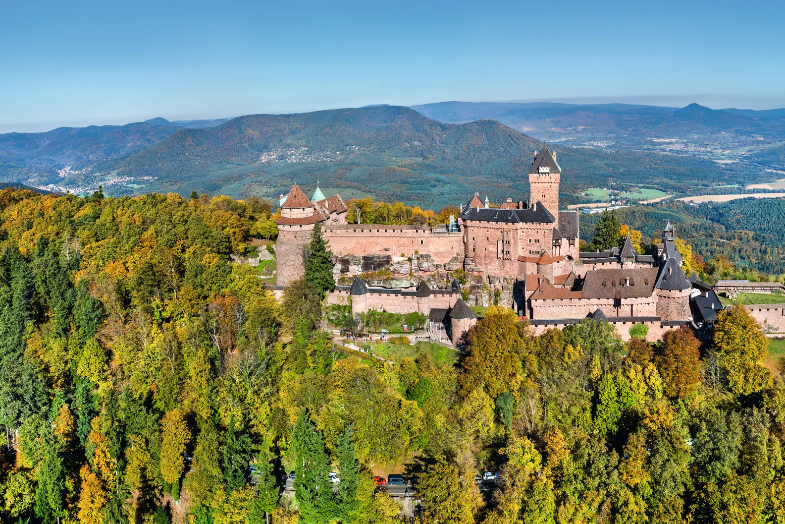Vue aérienne d’un grand château ancien sur une colline boisée dans un paysage automnal.