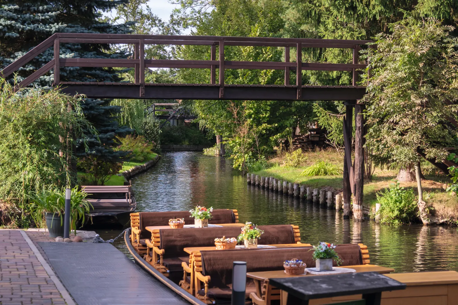 Bateaux d’excursion aux tables dressées sur une rivière idyllique, avec un pont en bois et une végétation luxuriante.