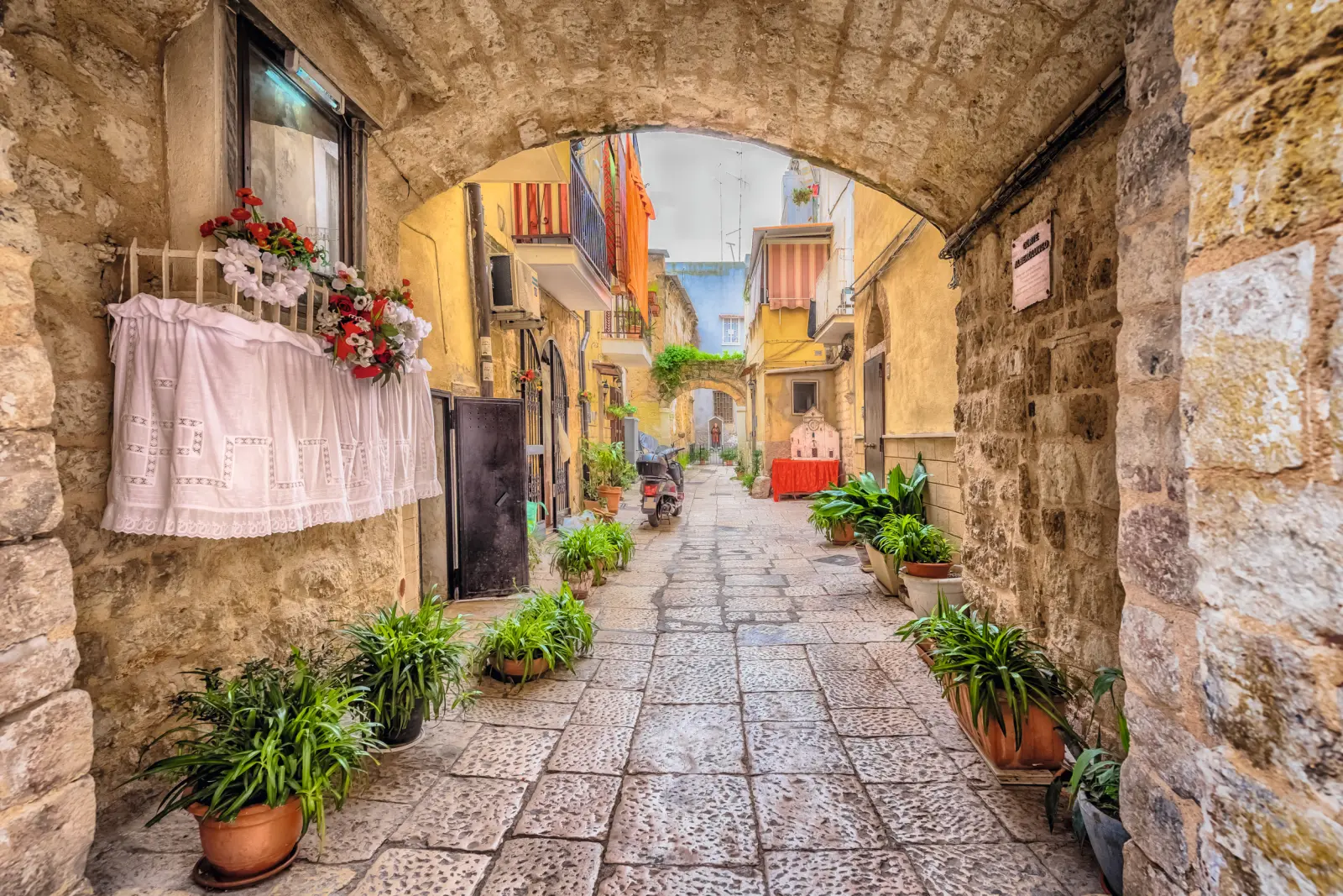Arco in pietra su un vicolo con piante ed edifici colorati in un villaggio mediterraneo.