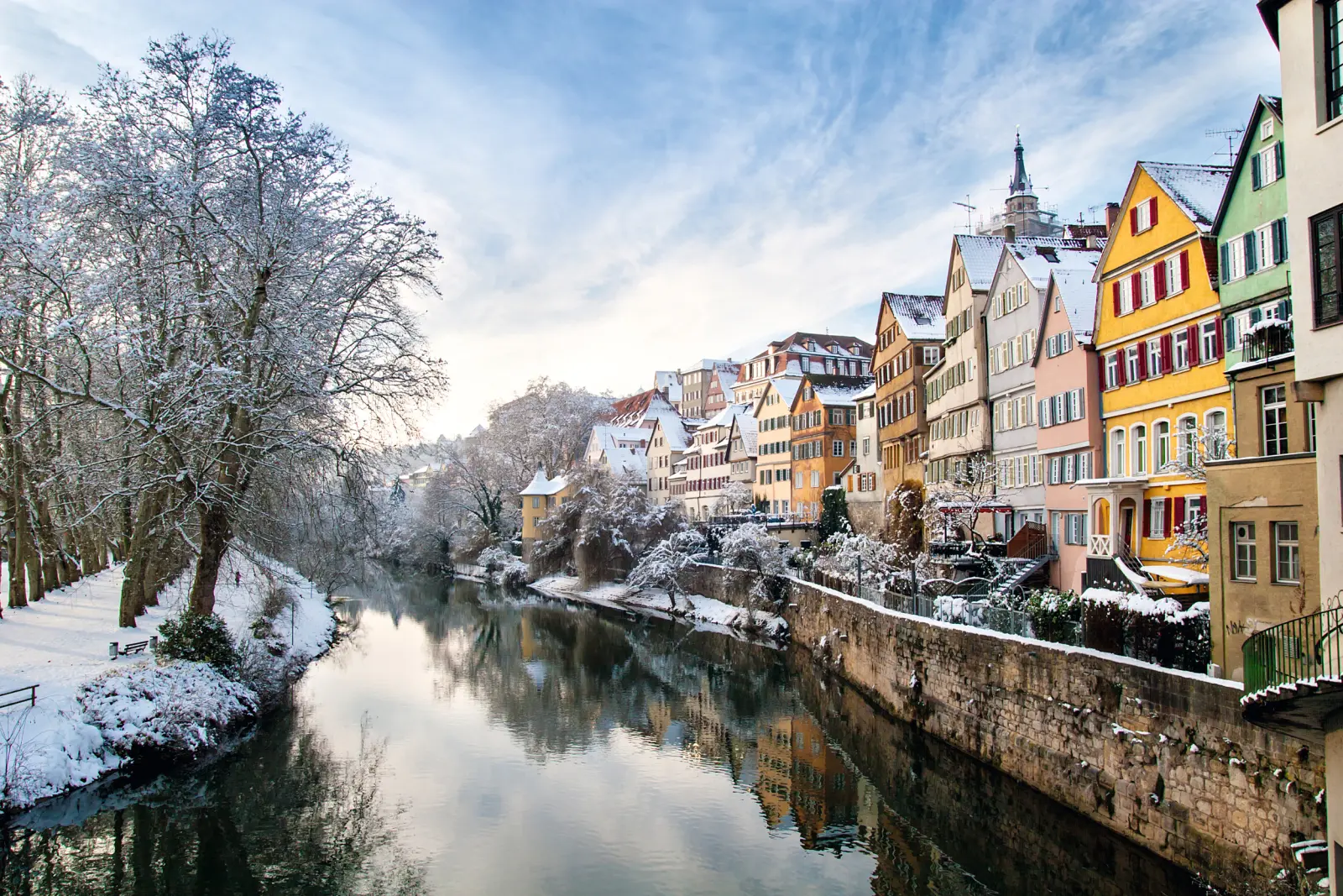 Vue d’une ville enneigée au bord d’une rivière, avec des maisons colorées et des arbres en hiver.
