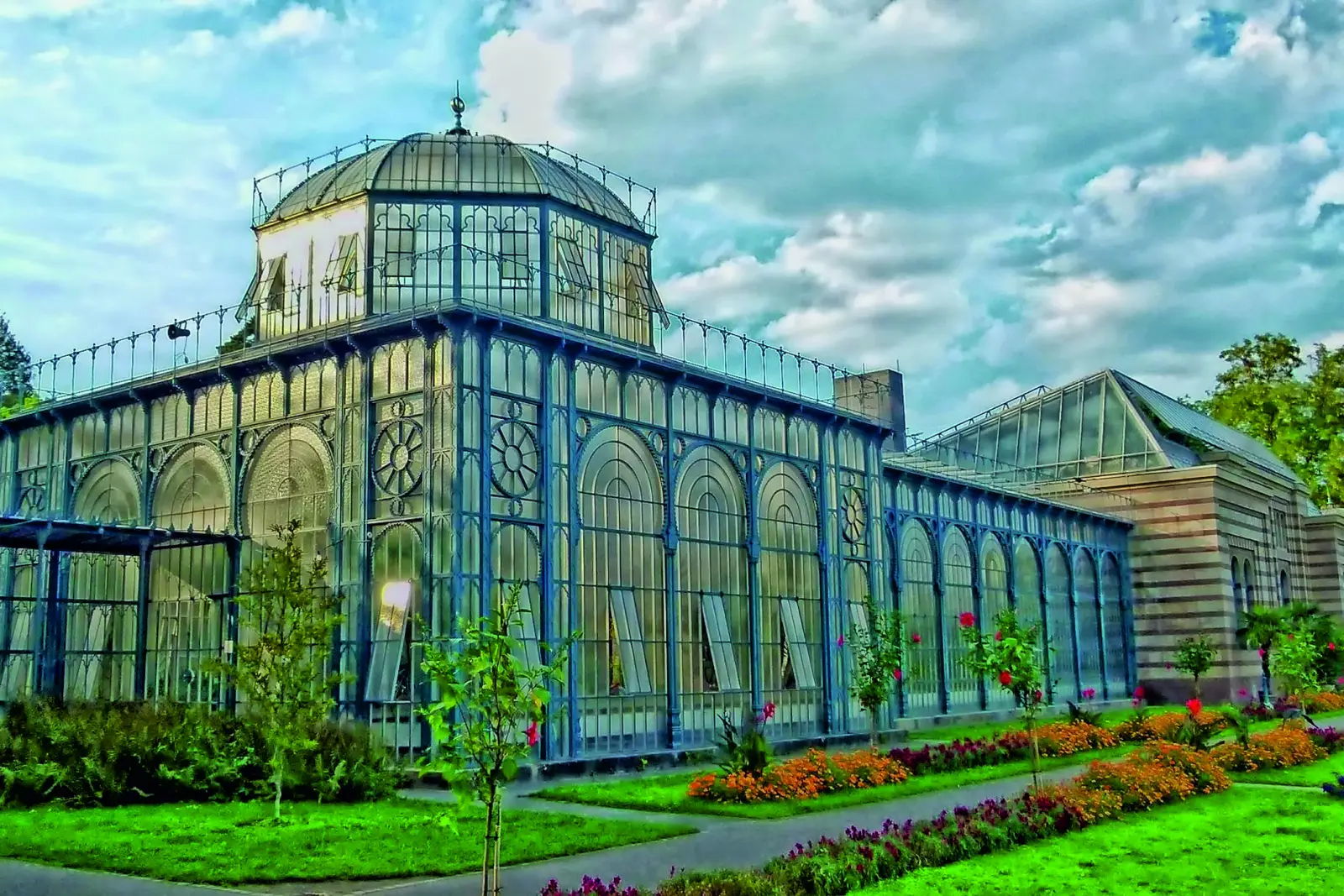 Historisches Gewächshaus mit dekorativer Fassade und blühenden Gärten im Vordergrund.