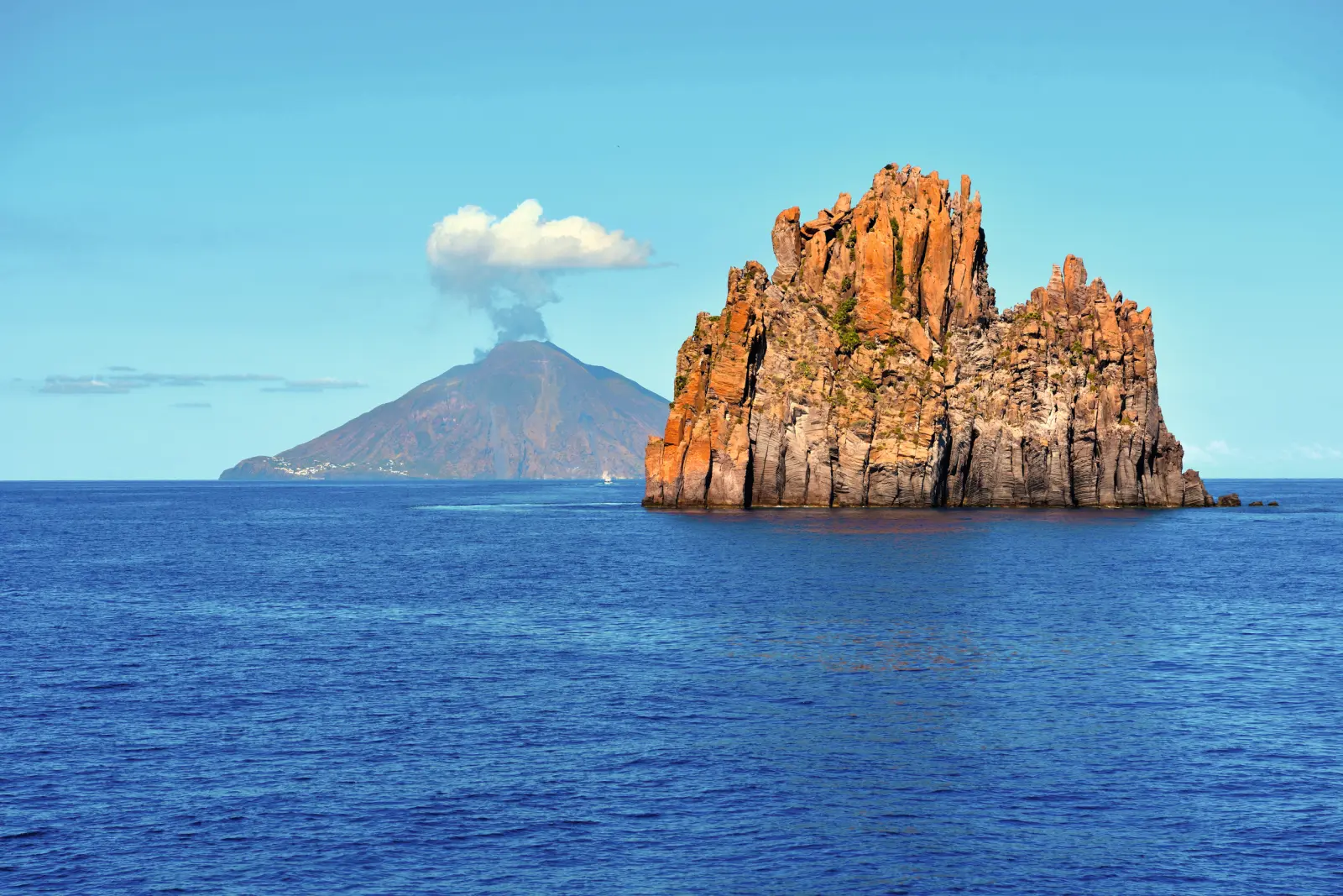 Formation rocheuse dans la mer avec un volcan fumant à l’arrière-plan sous un ciel bleu.