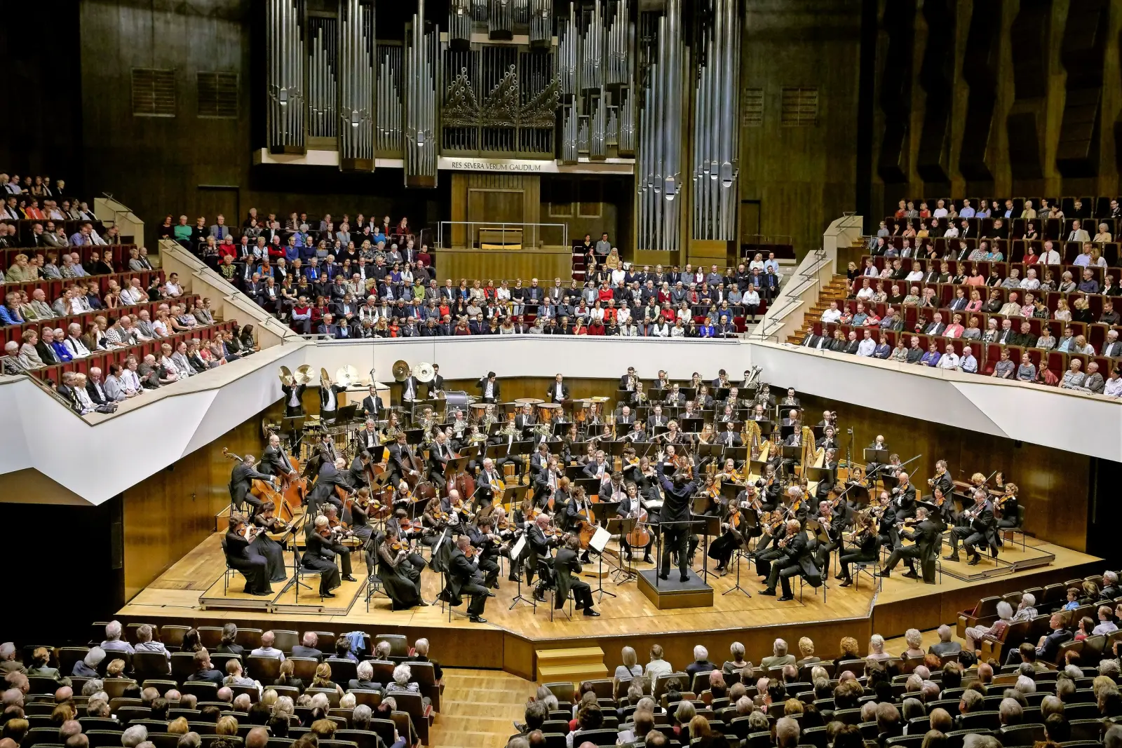 Ein Orchester spielt vor einem großen Publikum in einem Konzertsaal.