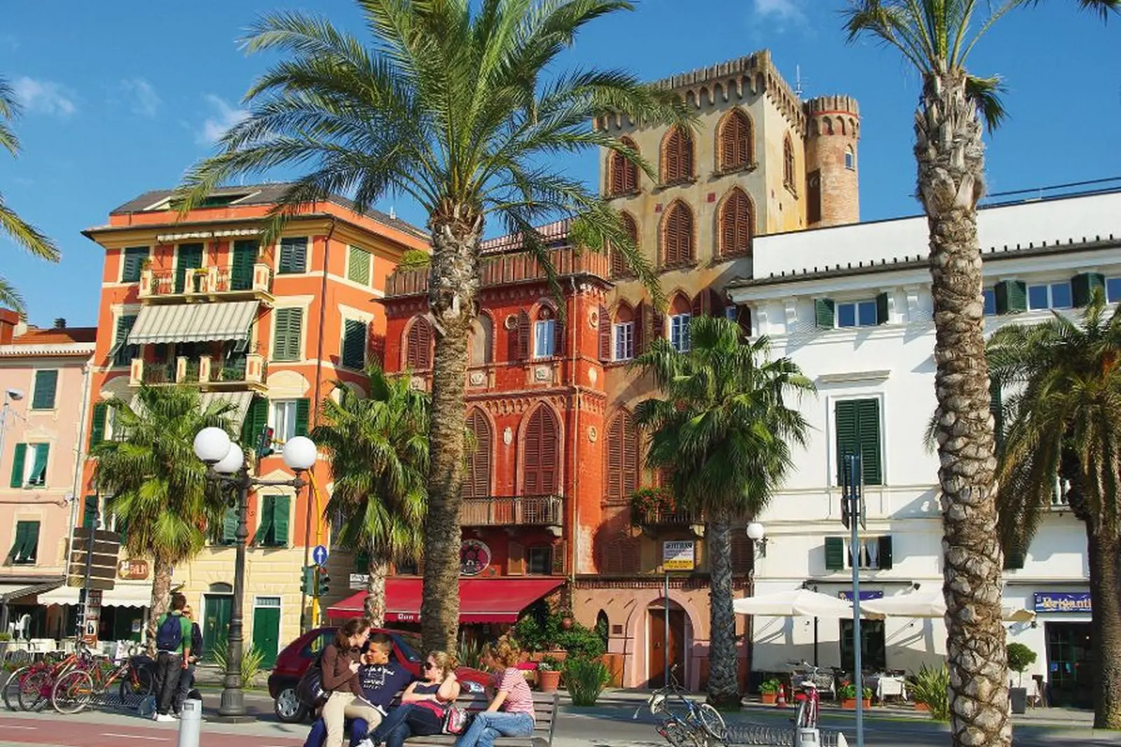 Colourful buildings with palm trees in a town, people sitting in the street.