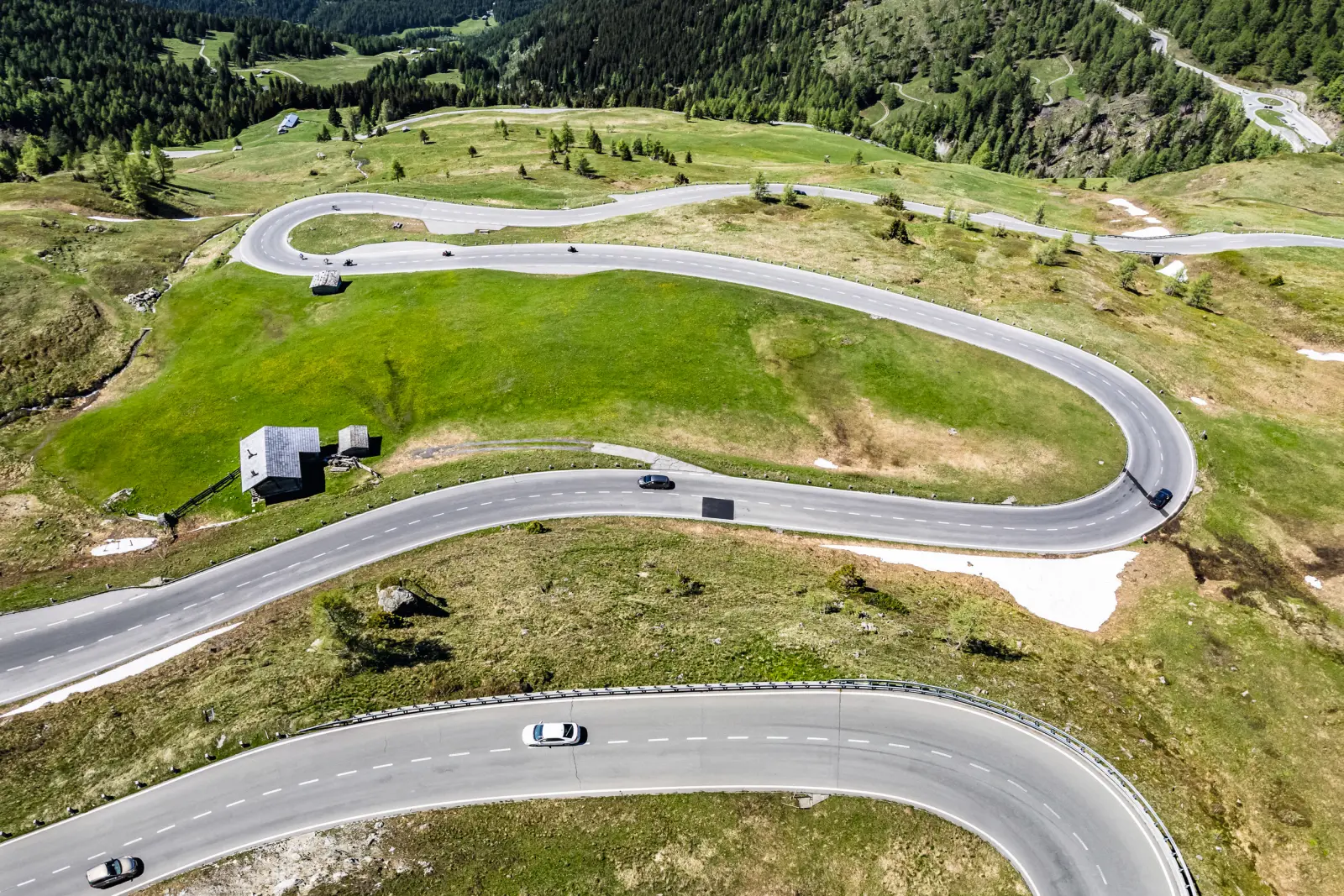 Winding mountain road with cars, green meadows and snow-capped mountains in the background.