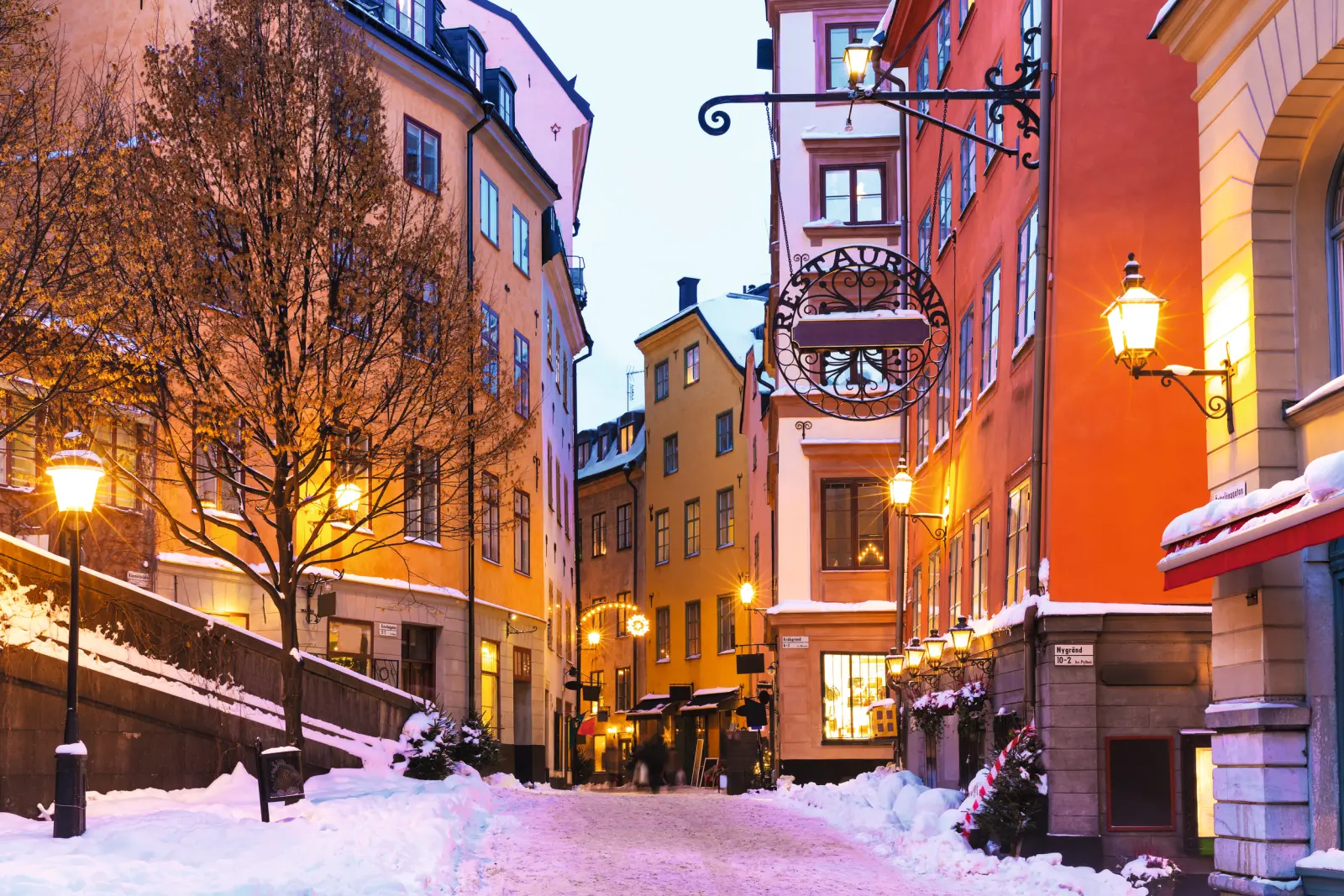 Vicolo innevato del centro storico con facciate colorate e lampioni al crepuscolo.