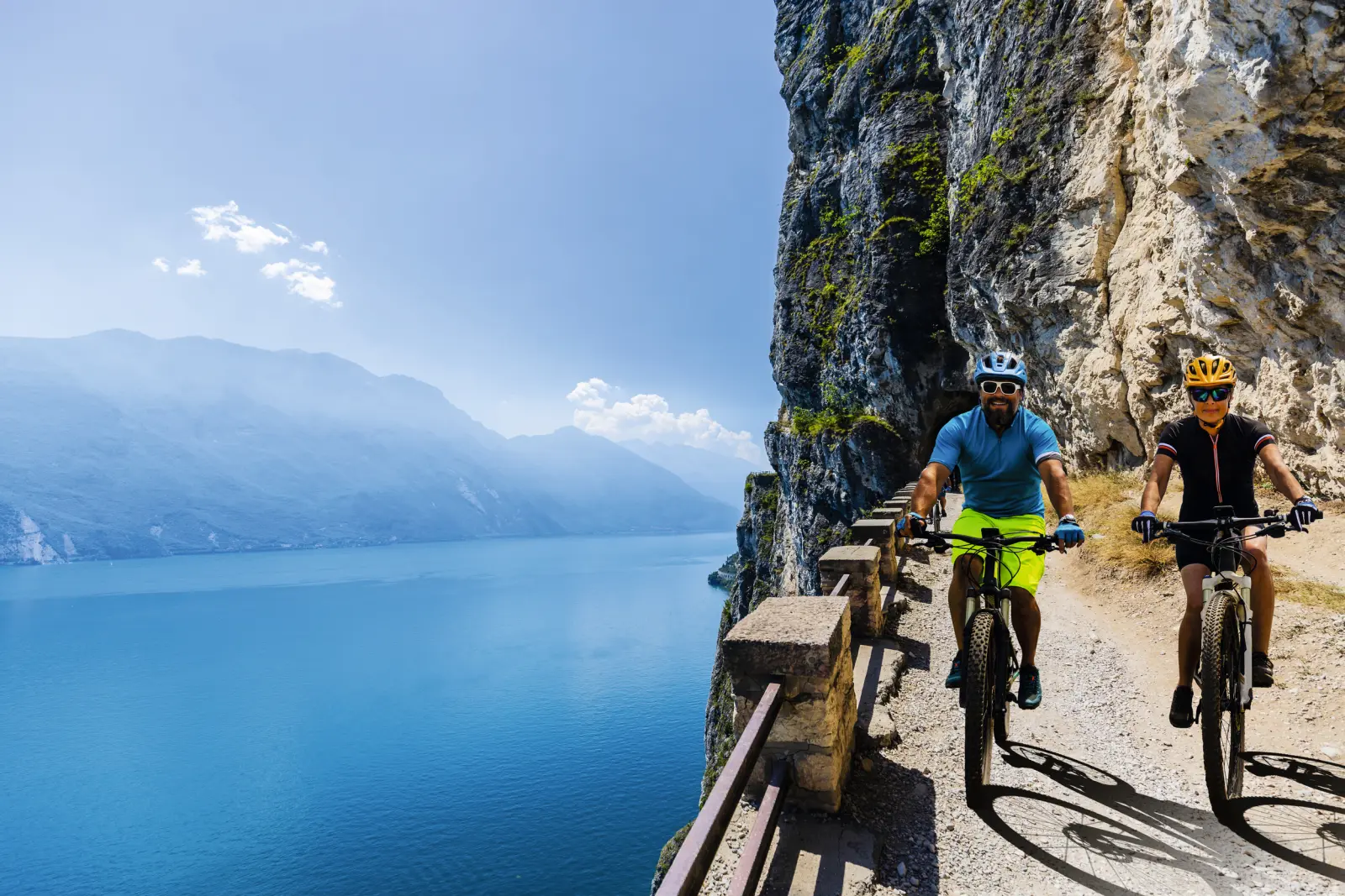 Zwei Radfahrer auf einem schmalen Weg entlang eines Sees und einer Felswand.
