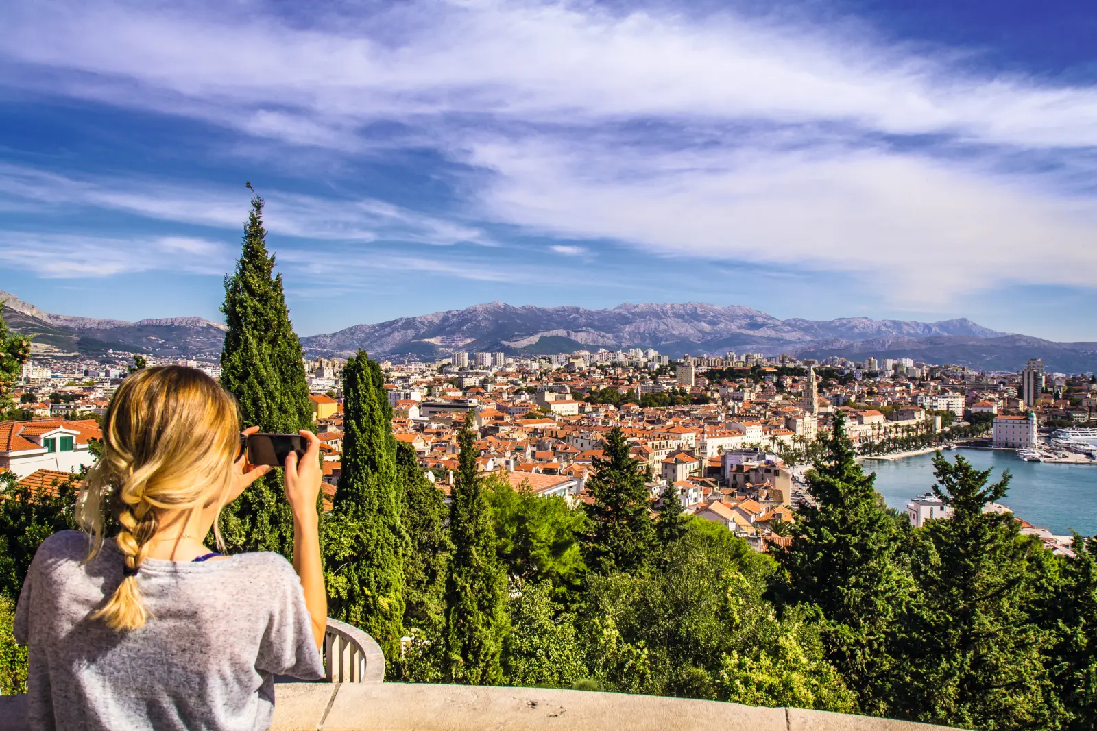 Femme photographiant la ville et la côte depuis un point de vue entouré d’arbres.