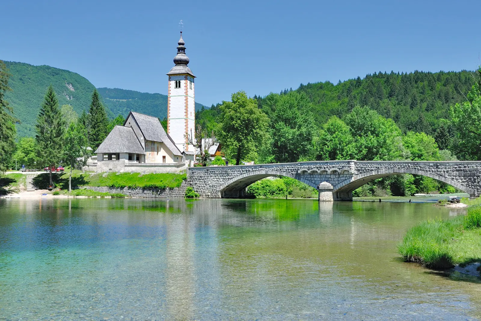 Kirche neben steinernem Brücke am See vor bewaldeten Hügeln bei klarem Himmel.