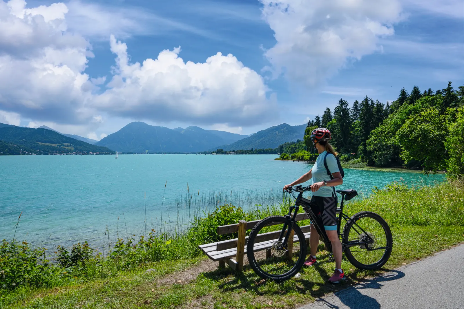 Person mit Fahrrad am See mit Bergen im Hintergrund.
