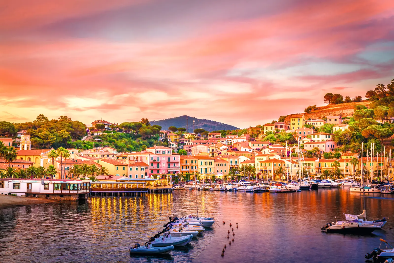 Harbour of Porto Azzurro on the island of Elba at sunset, with boats on the water and colourful buildings in the background.