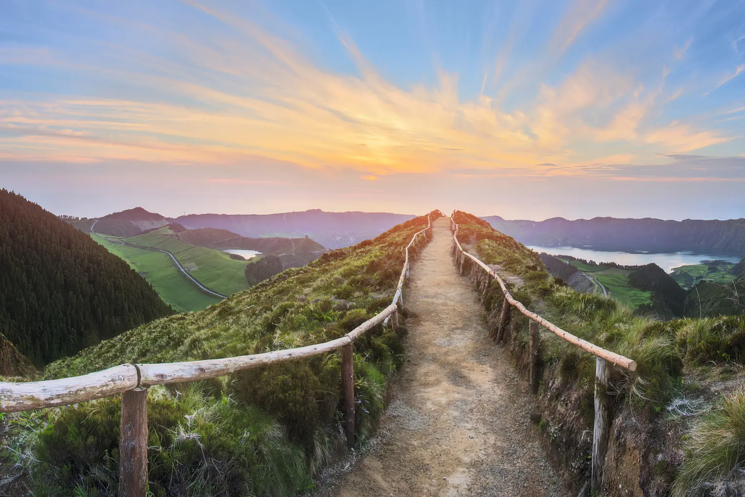 Path with a wooden railing leading through green, hilly countryside at sunset.