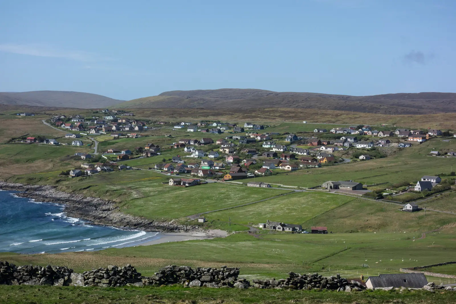 Coastal landscape with a village and green hills under a blue sky.