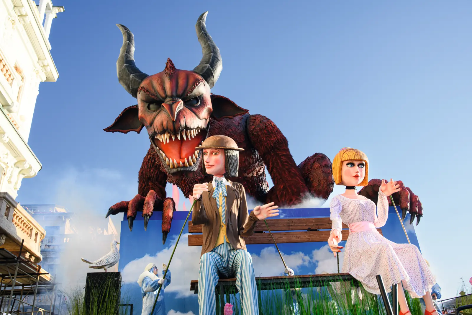 Carnival float with a huge devil figure and two puppets against a blue sky.