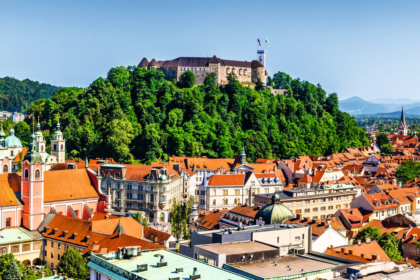 Altstadt von Ljubljana mit der mittelalterlichen Burg auf einem bewaldeten Hügel im Hintergrund.