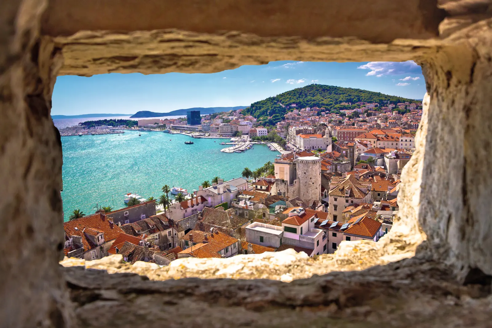 Vue à travers une fenêtre en pierre sur une ville côtière, avec la mer et une colline à l’arrière-plan.