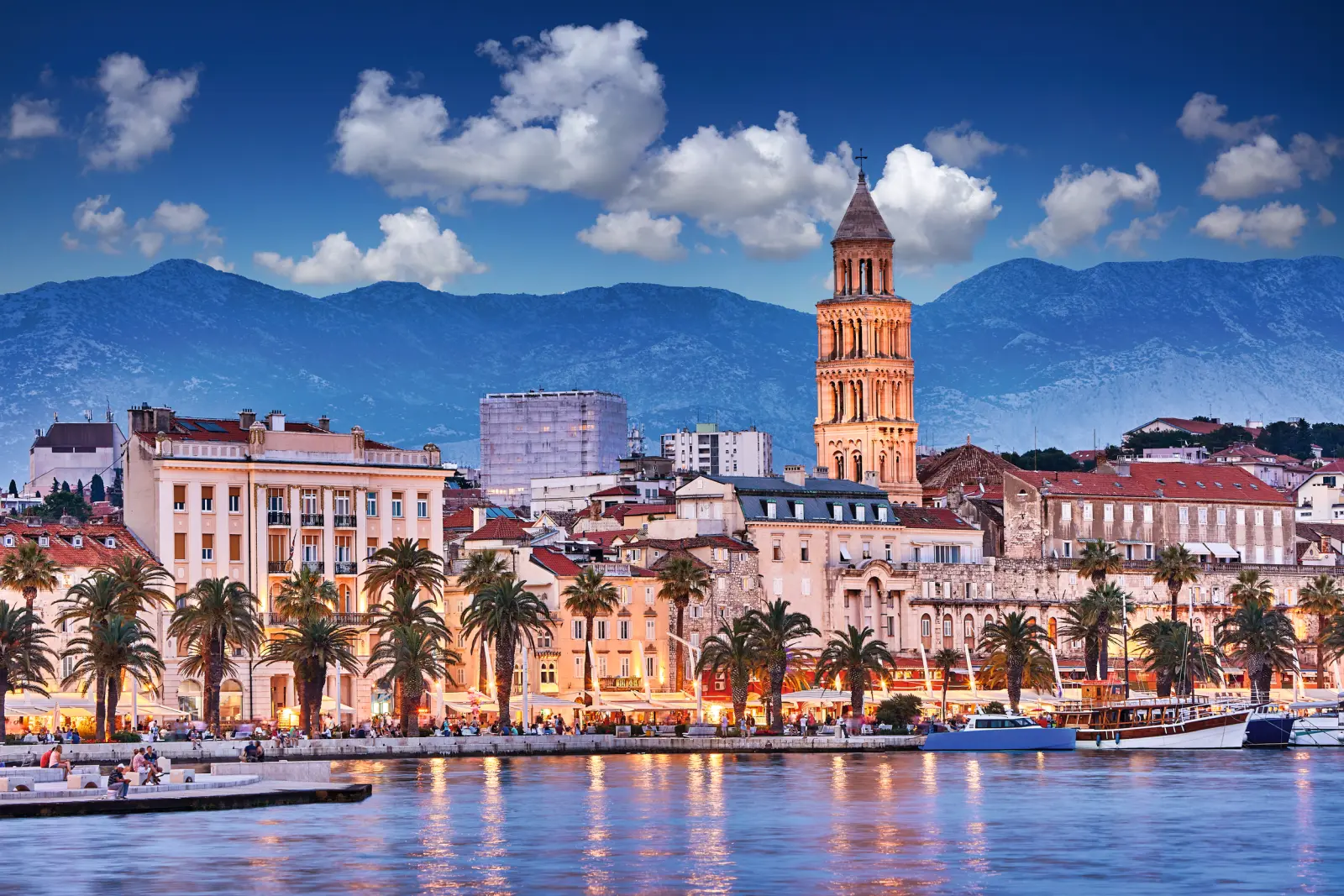 Città storica sul mare con montagne e torre illuminata al crepuscolo.
