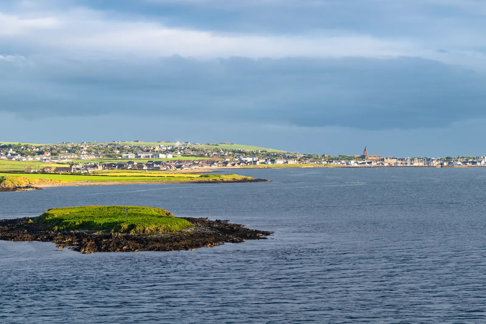 Island in the sea with a town in the background under a cloudy sky