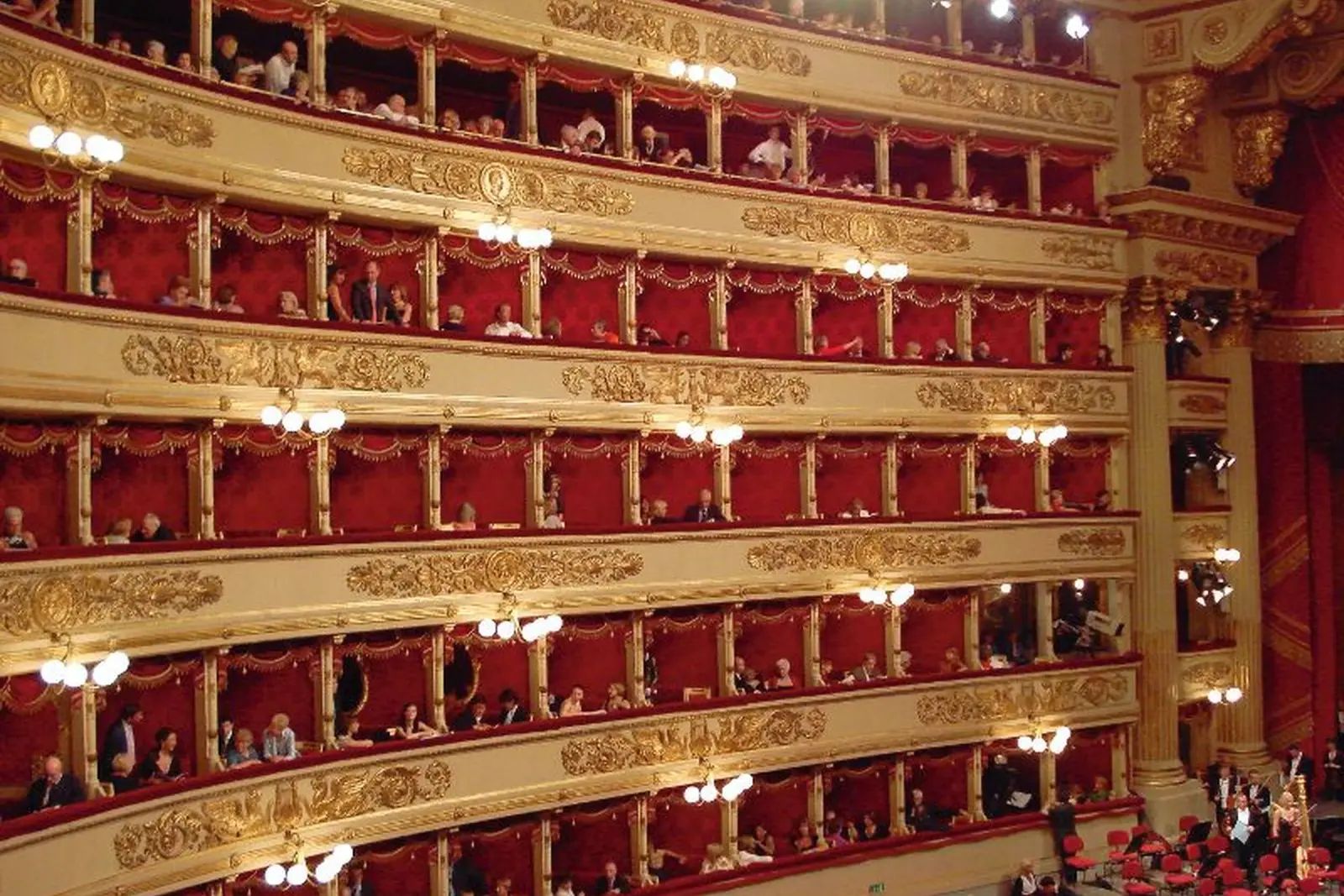 Veduta interna di un teatro con palchi rossi e spettatori.