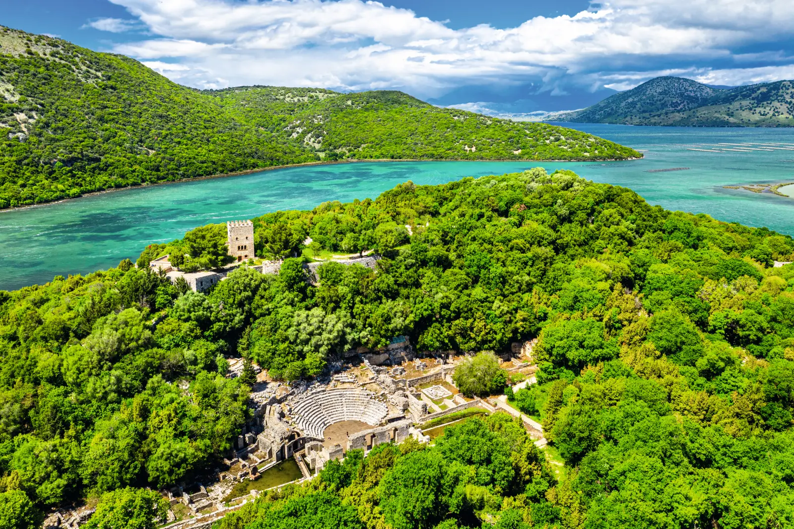 Aerial view of an ancient theatre amid green hills beside a turquoise lake.