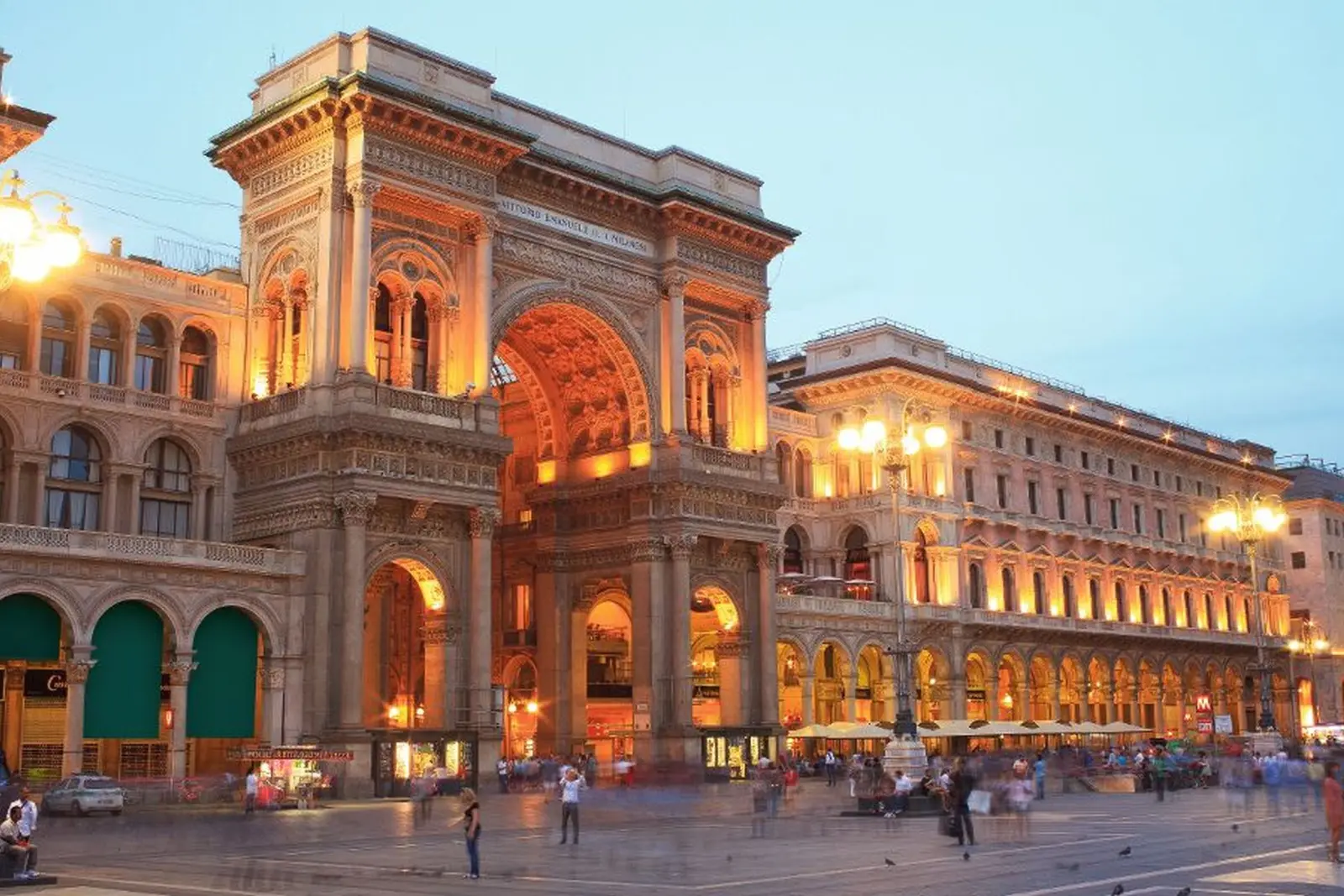 Bâtiment historique en soirée, avec arcades illuminées et place animée.