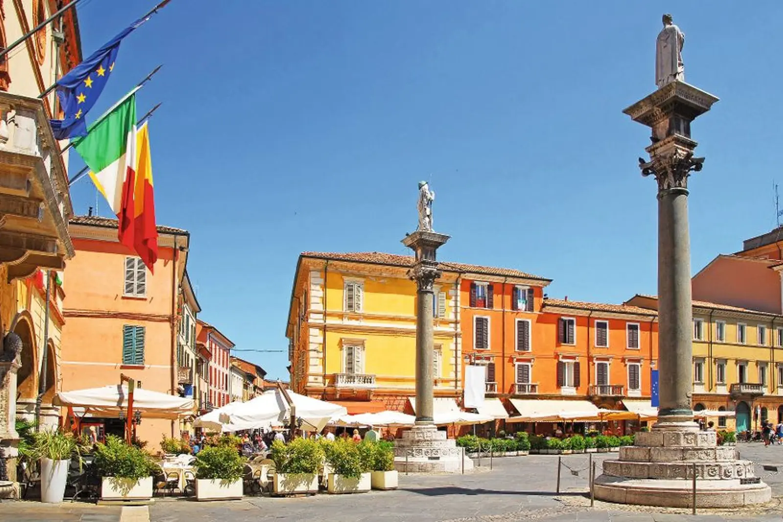 Due alte colonne di pietra con statue, circondate da edifici colorati e bandiere al vento.