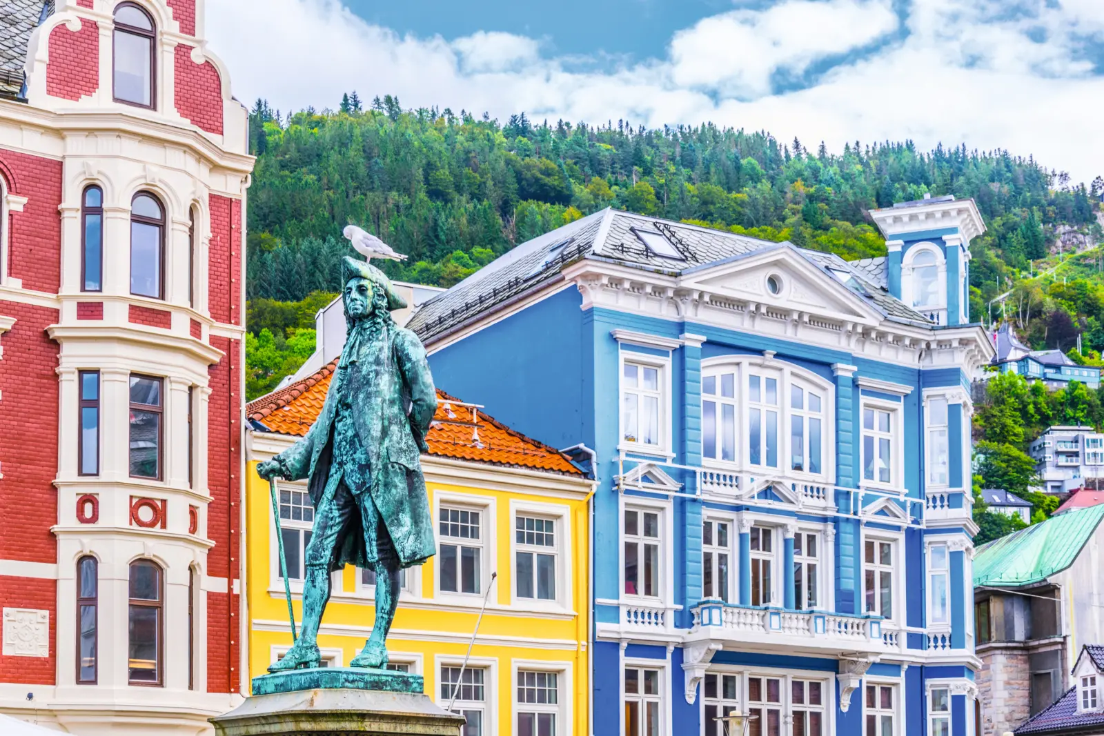 Statue mit Möwe, bunte Gebäude und bewaldeter Hügel im Hintergrund.