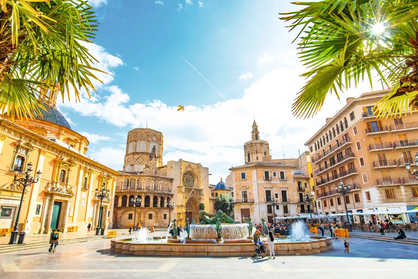 Historic town square with a fountain and surrounding buildings in the sunshine.