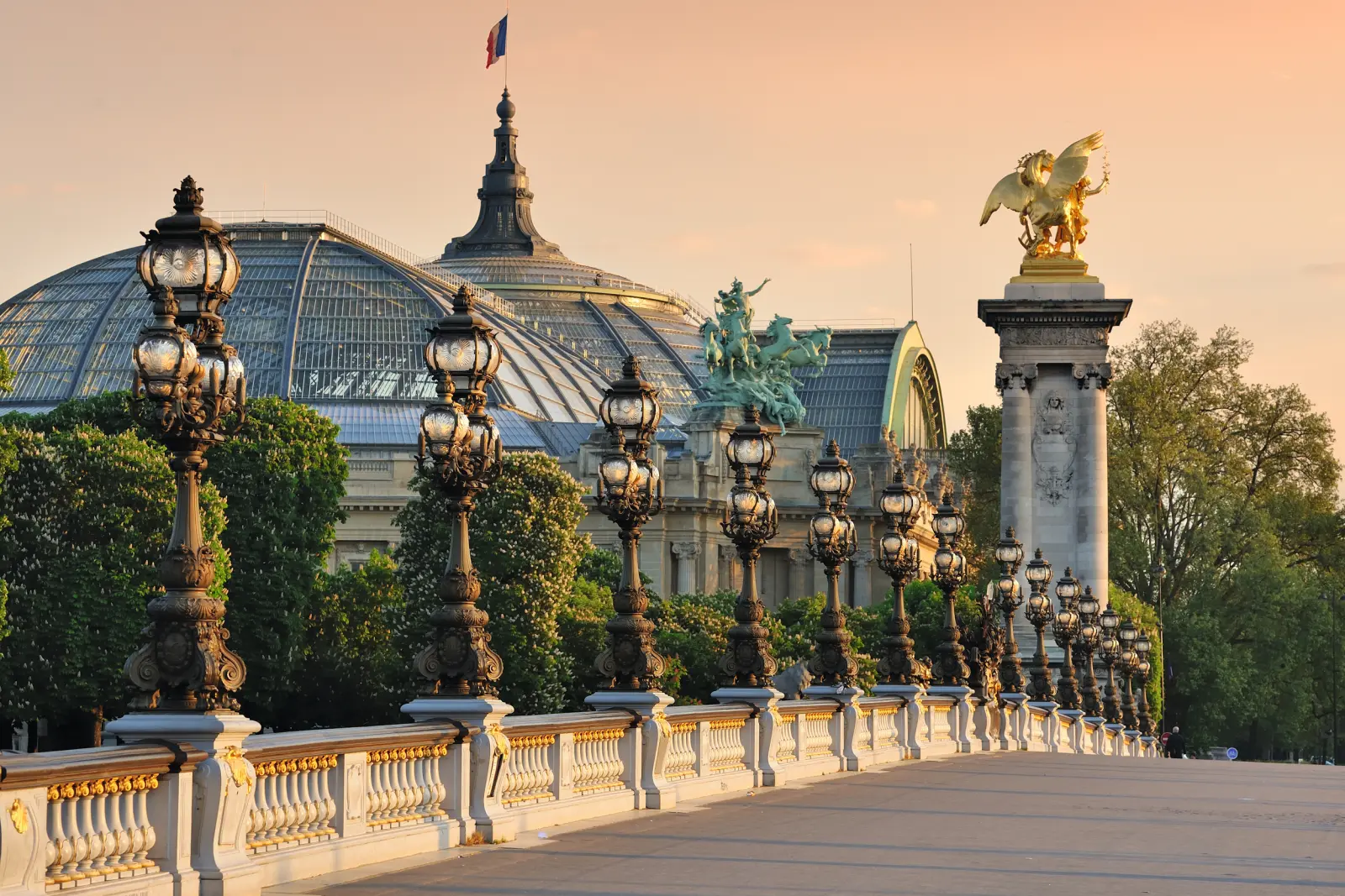 Vue sur le pont Alexandre III à Paris au coucher du soleil, avec le Grand Palais en arrière-plan.
