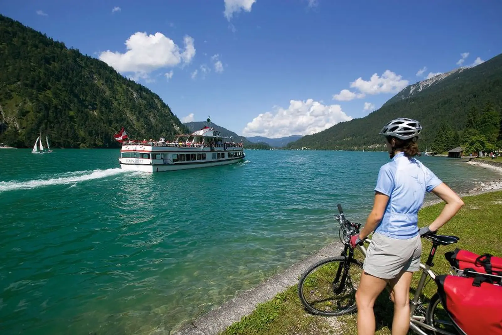 Frau mit Fahrrad am Ufer des Achensees, im Hintergrund ein Ausflugsschiff.