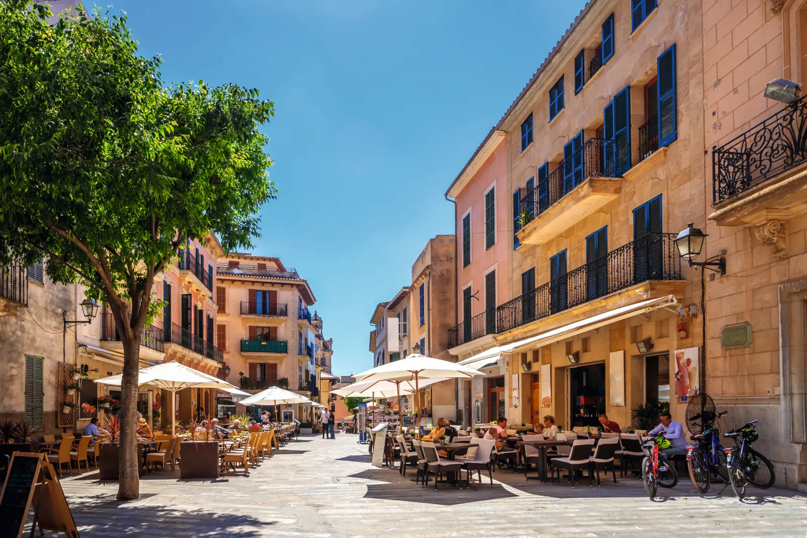 Caffè all'aperto in un soleggiato centro storico europeo con persone e biciclette.