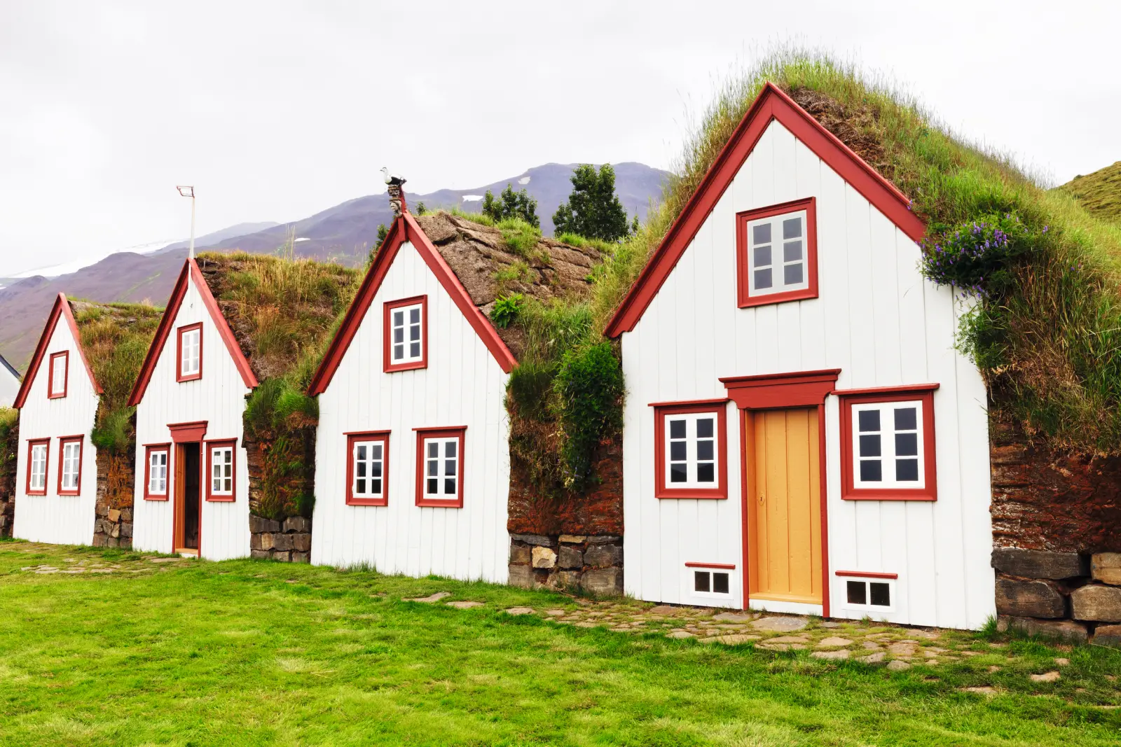 Three white wooden houses with grass roofs in a mountain landscape.