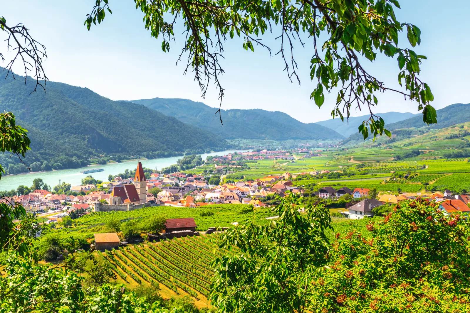 Vista panoramica su un villaggio con fiume e montagne circostanti sotto un cielo limpido.