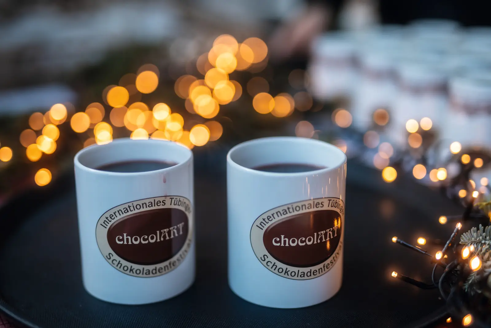 Two cups with the chocolART logo on a tray, with lights and more cups in the background.