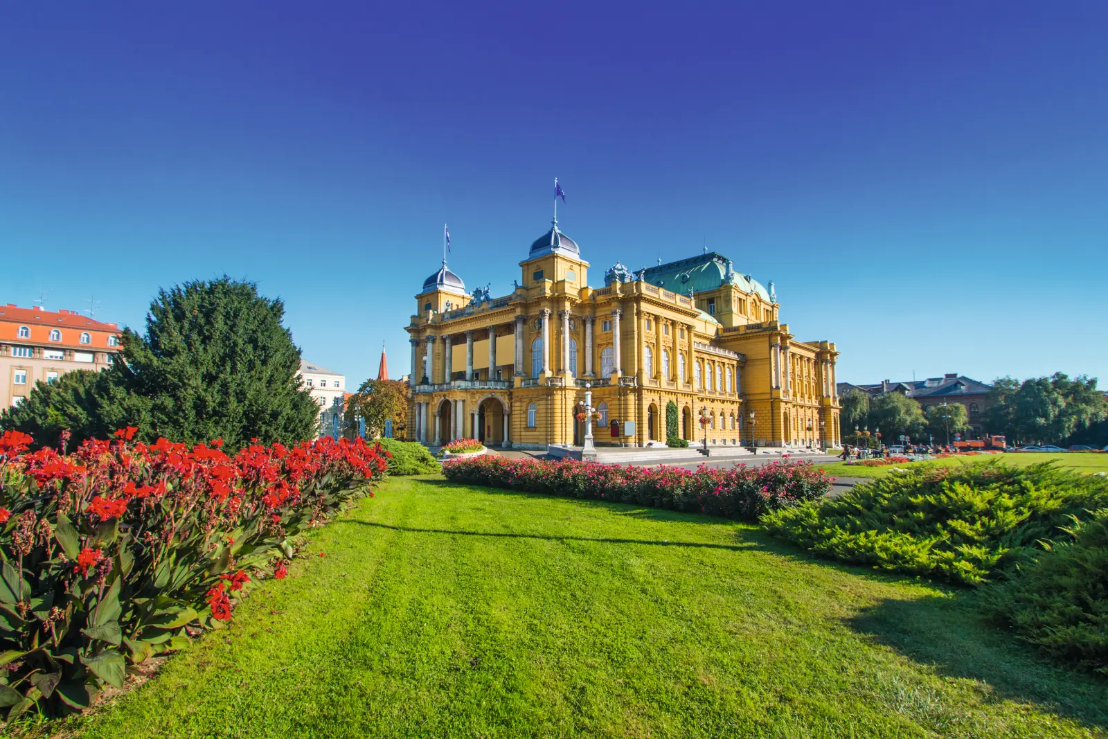 Edificio storico con parco, cielo azzurro e fiori rossi in primo piano a sinistra.