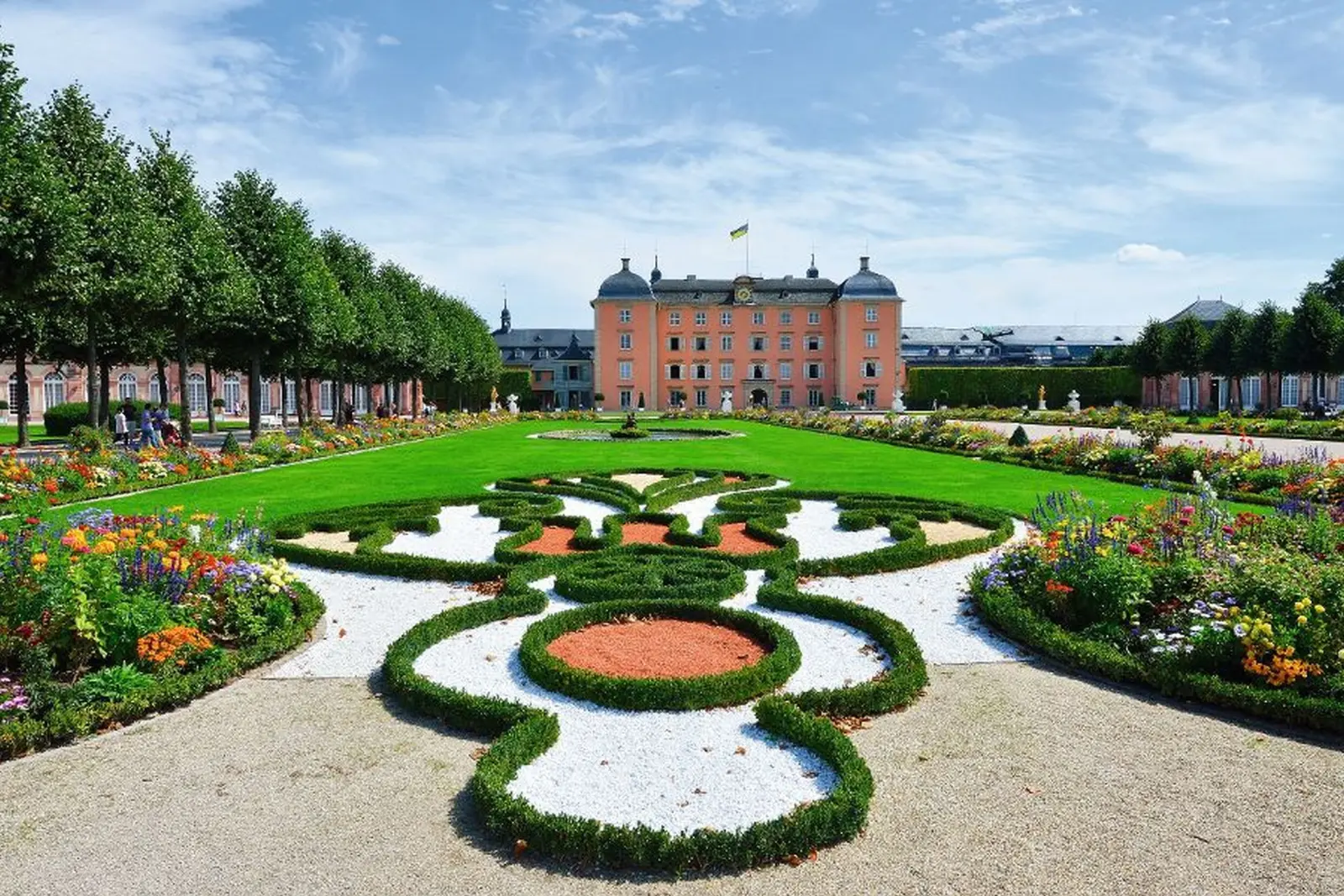 Jardin baroque devant un château rose sous un ciel dégagé.