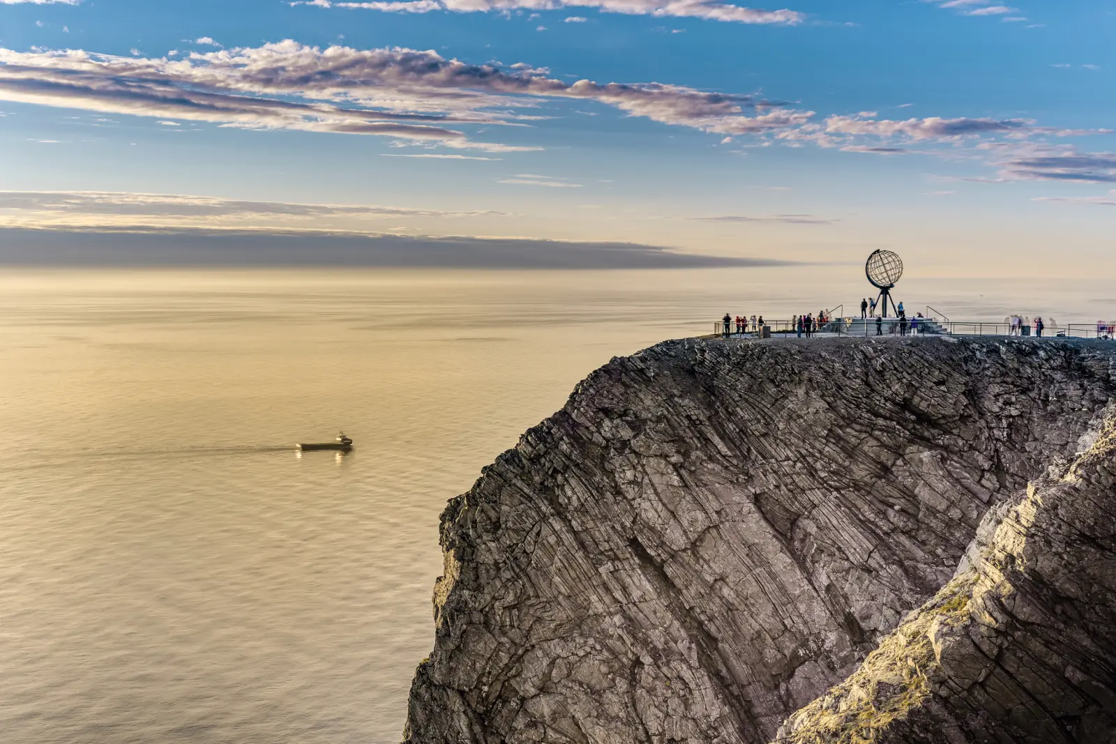 Rocky cliff with globe sculpture by the sea, people looking out over the water.