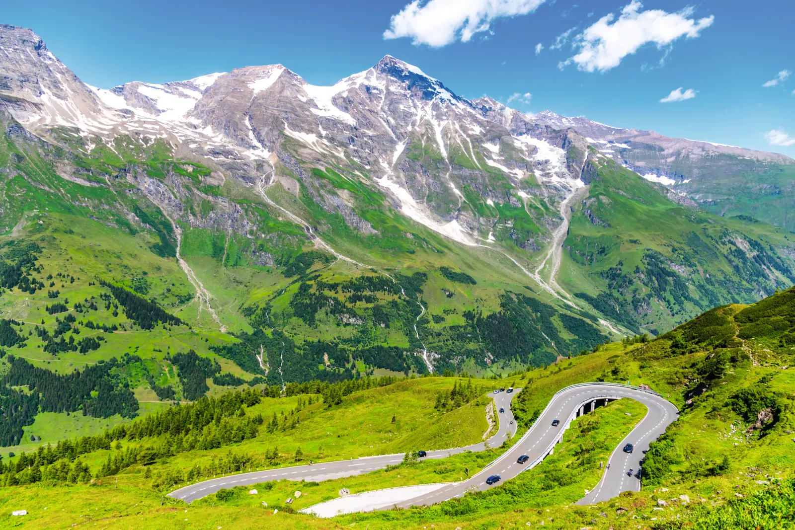 Strada a tornanti davanti a un paesaggio montano con neve e una valle verde.