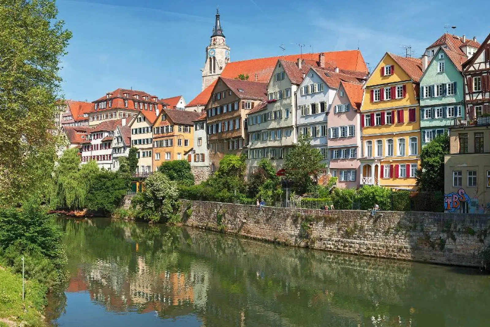Historic row of colourful houses on the riverbank with a church tower in the background.