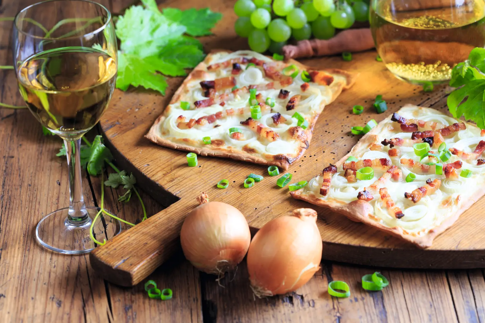 Tarte flambée aux lardons et aux oignons sur une planche en bois, accompagnée de vin blanc et de raisins.