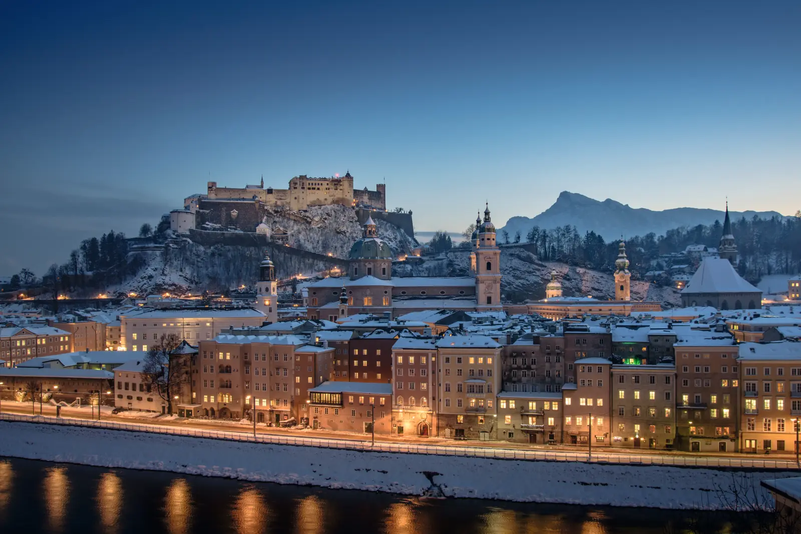 Città innevata con castello al crepuscolo, illuminati lungo un fiume.