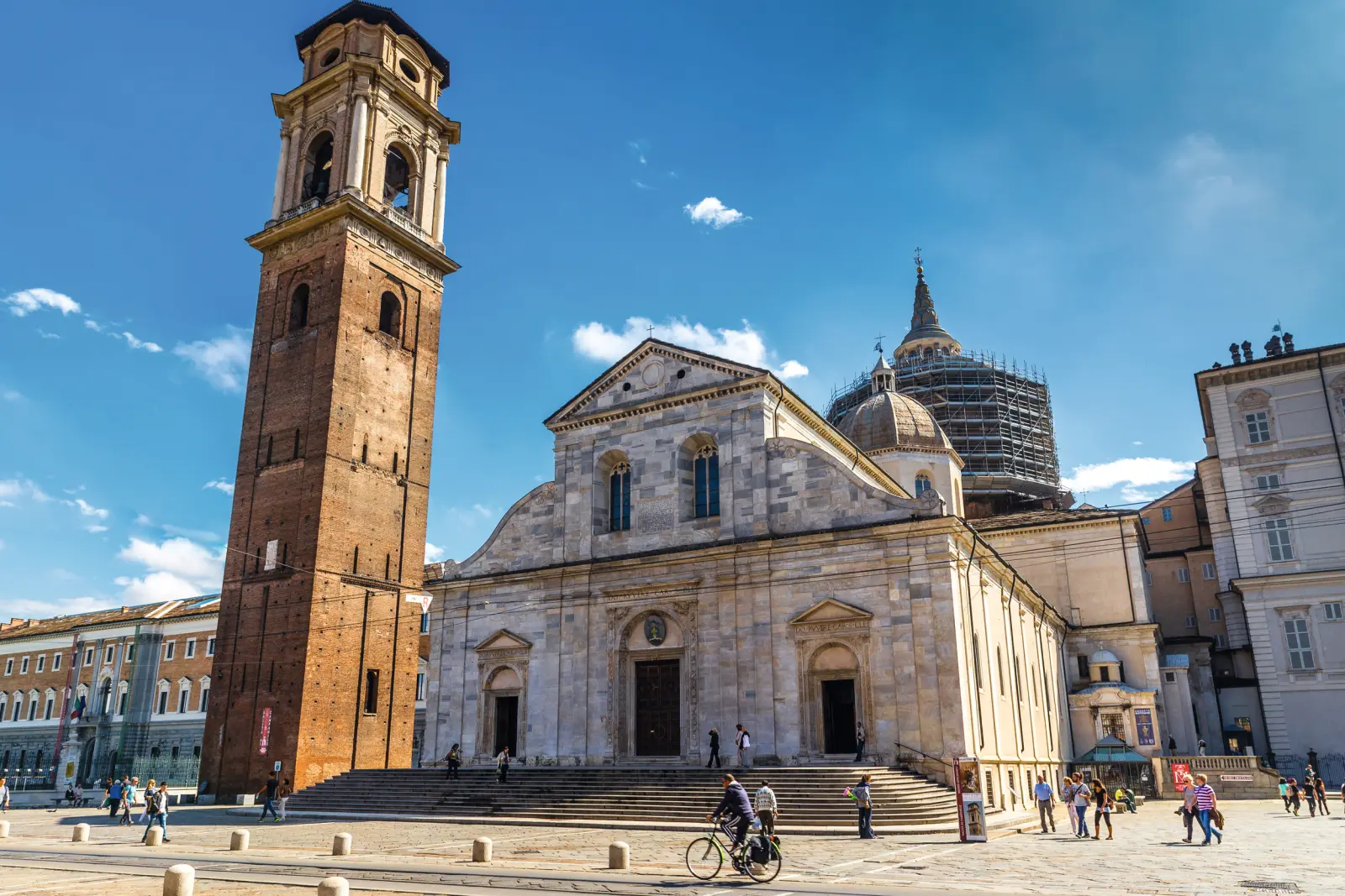 Cathédrale et clocher sur une place ensoleillée, des gens se promènent à pied et à vélo.