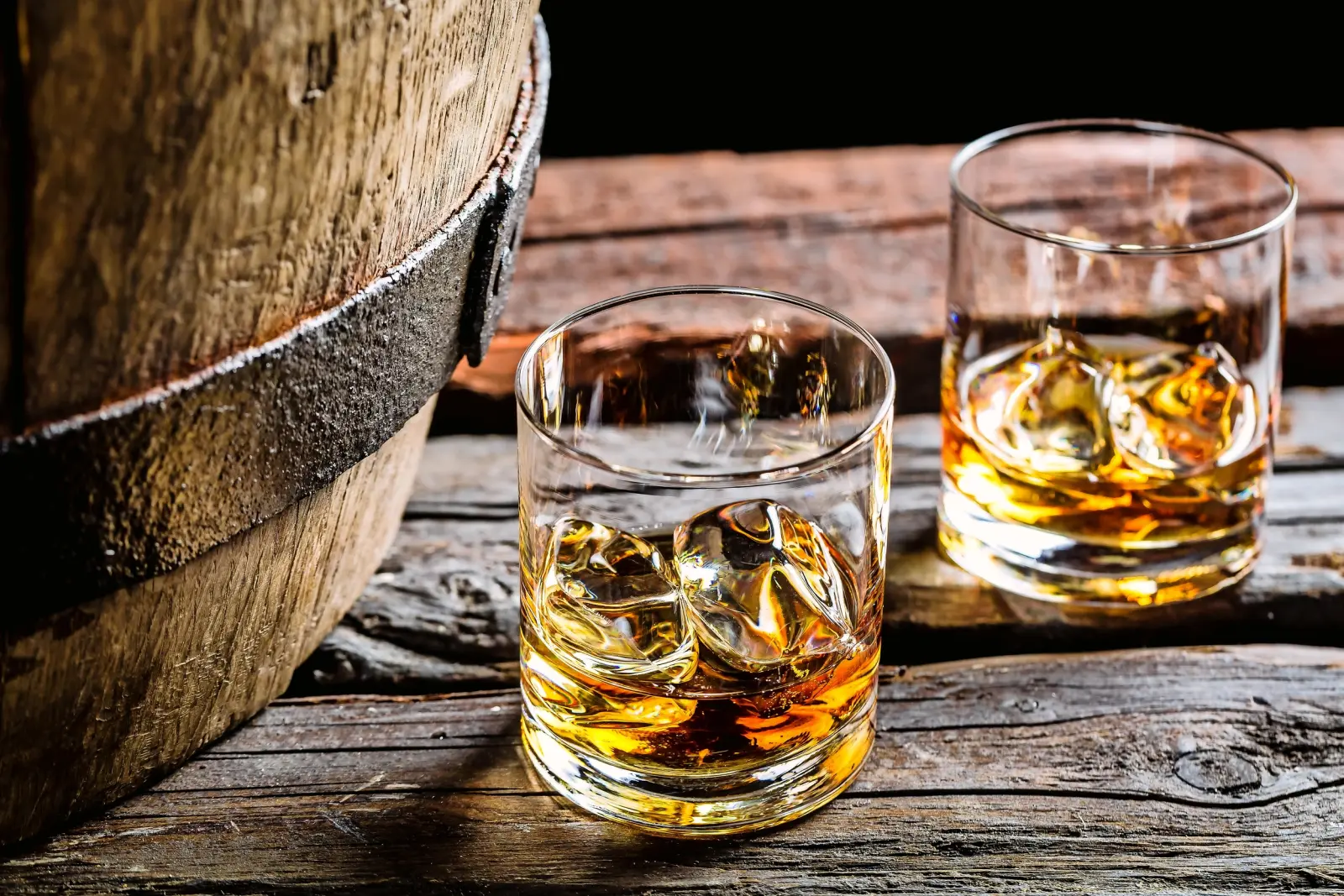 Deux verres de whisky avec des glaçons sur une table en bois à côté d’un tonneau.
