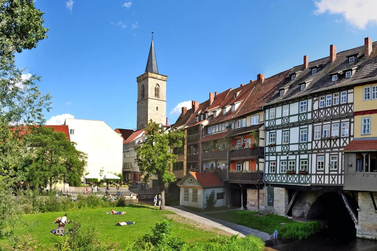 Pont avec maisons à colombages et clocher d’église, parc verdoyant au premier plan.