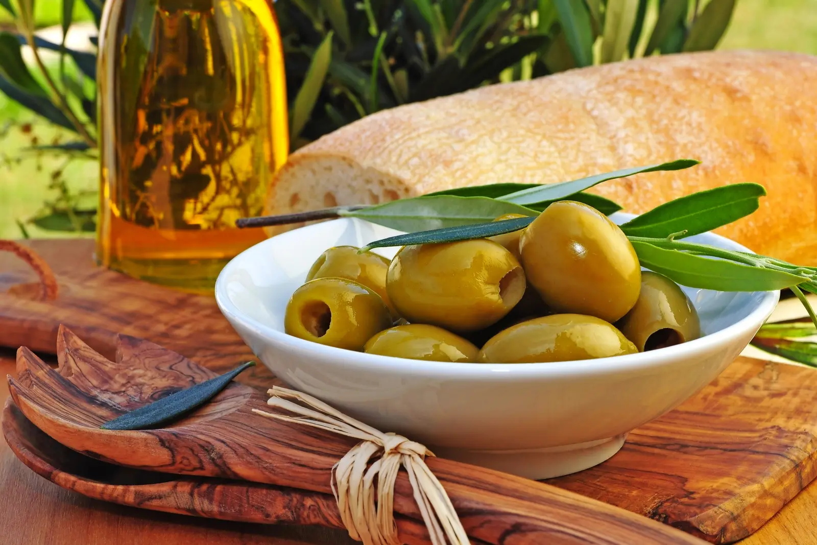 Schale mit grünen Oliven, Olivenölflasche, Holzlöffel, frisches Brot im Hintergrund.
